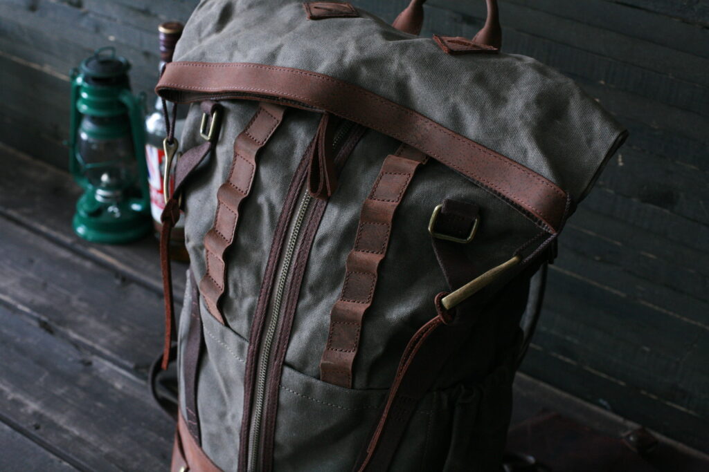 Premium Heritage Gear: Builford Bikers Crew Backpack Detail A front view of the Builford Bikers Crew Backpack in olive waxed canvas and brown leather, placed on a dark wooden floor next to a vintage lantern.