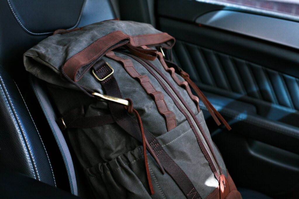 Premium Versatility: Builford Bikers Crew Backpack in Daily Life A Builford Bikers Crew Backpack in olive waxed canvas resting on a black leather car seat, highlighting its premium texture and heritage design.