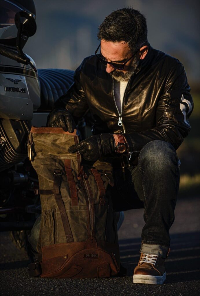 Stylish Commuting: Builford Bikers Crew Backpack for Modern Riders A mature male rider in a black leather jacket and sunglasses examining a Builford Bikers Crew Backpack next to his motorcycle.