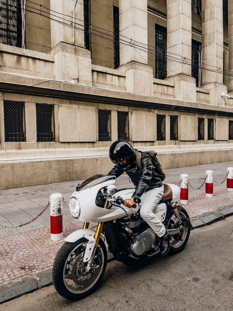 Urban Heritage: Builford Bikers Crew Backpack for the Modern City Rider A rider on a white classic motorcycle cruising through a city street with historic architecture, wearing a Builford Bikers Crew Backpack.